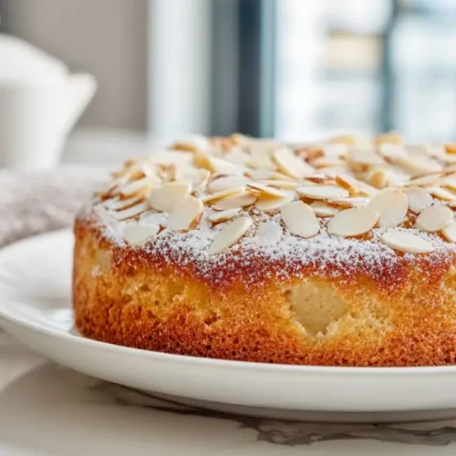 Golden apple almond cake topped with sliced almonds and powdered sugar on a white plate with teapot in background