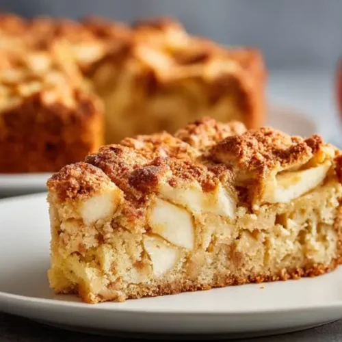 slice of apple crumb cake with soft apple chunks and golden streusel topping on plate with apples in background