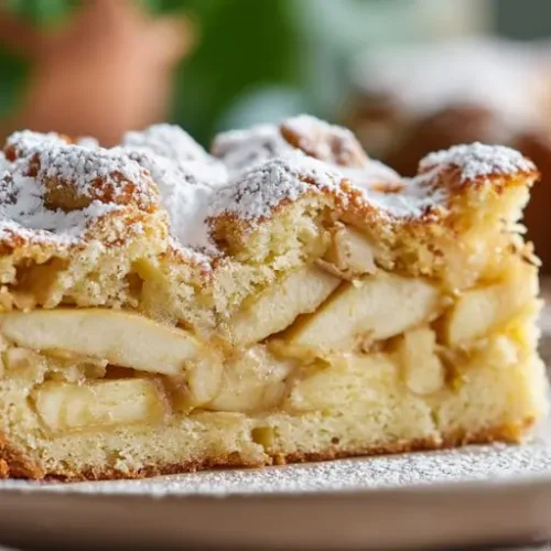 Moist Jewish apple cake slice filled with cinnamon-spiced apple layers and topped with powdered sugar on a dessert plate.