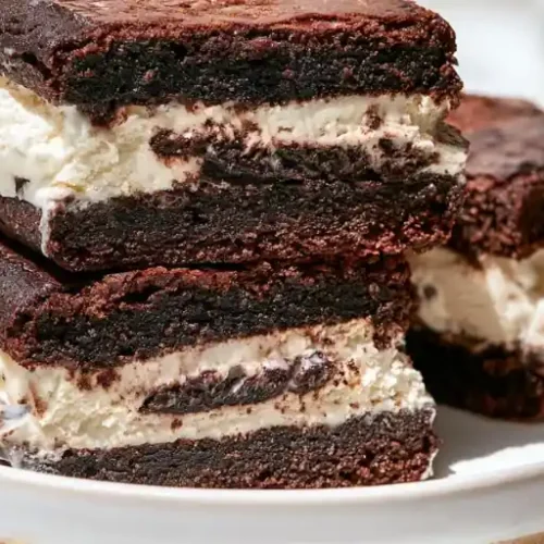 Stacked fudgy chocolate brownie sandwiches filled with creamy vanilla frosting on a white plate.